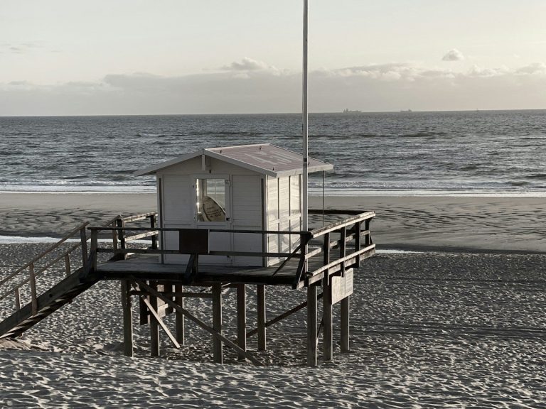 Badeaufsichtshütte auf Holzplattform am Strand mit Wellen im Hintergrund.