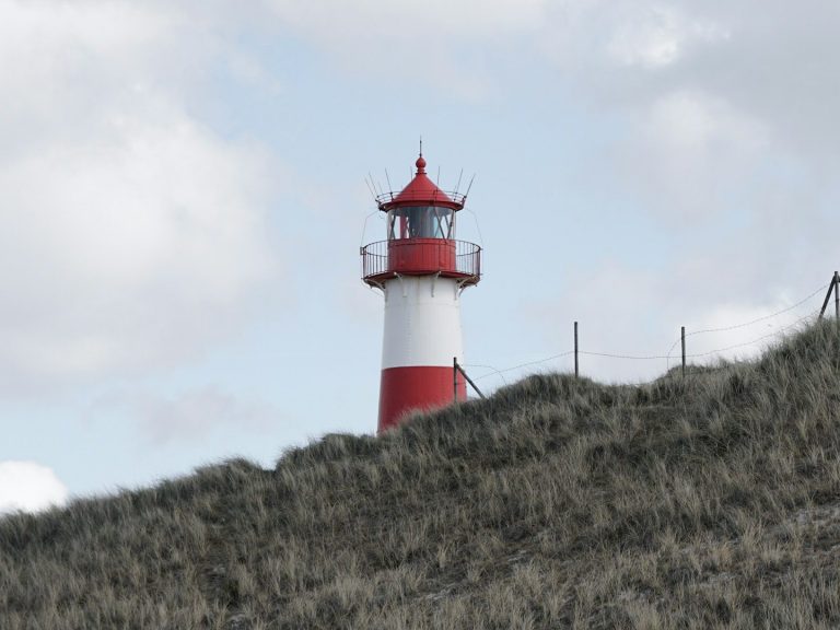 Leuchtturm mit rotem Dach, umgeben von Dünen und bewölktem Himmel.